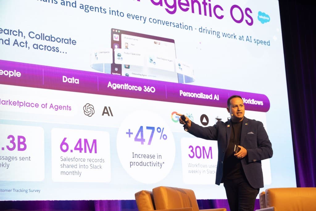 Presenter on a large conference stage in front of a screen showing Salesforce and Agentic OS narrative