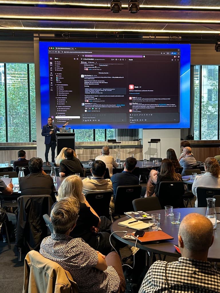 Presenter at a lectern with a projected Slack workspace and Claude AI assistant panel